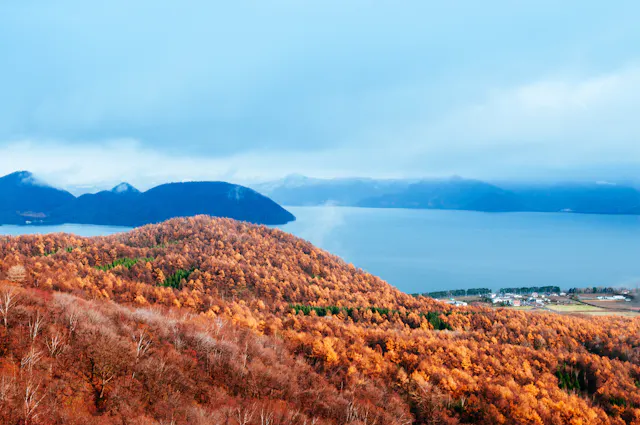 A scenic view of a lake surrounded by rolling hills covered in autumn trees with orange and brown leaves, distant mountains under a cloudy blue sky, and a small village near the water’s edge.