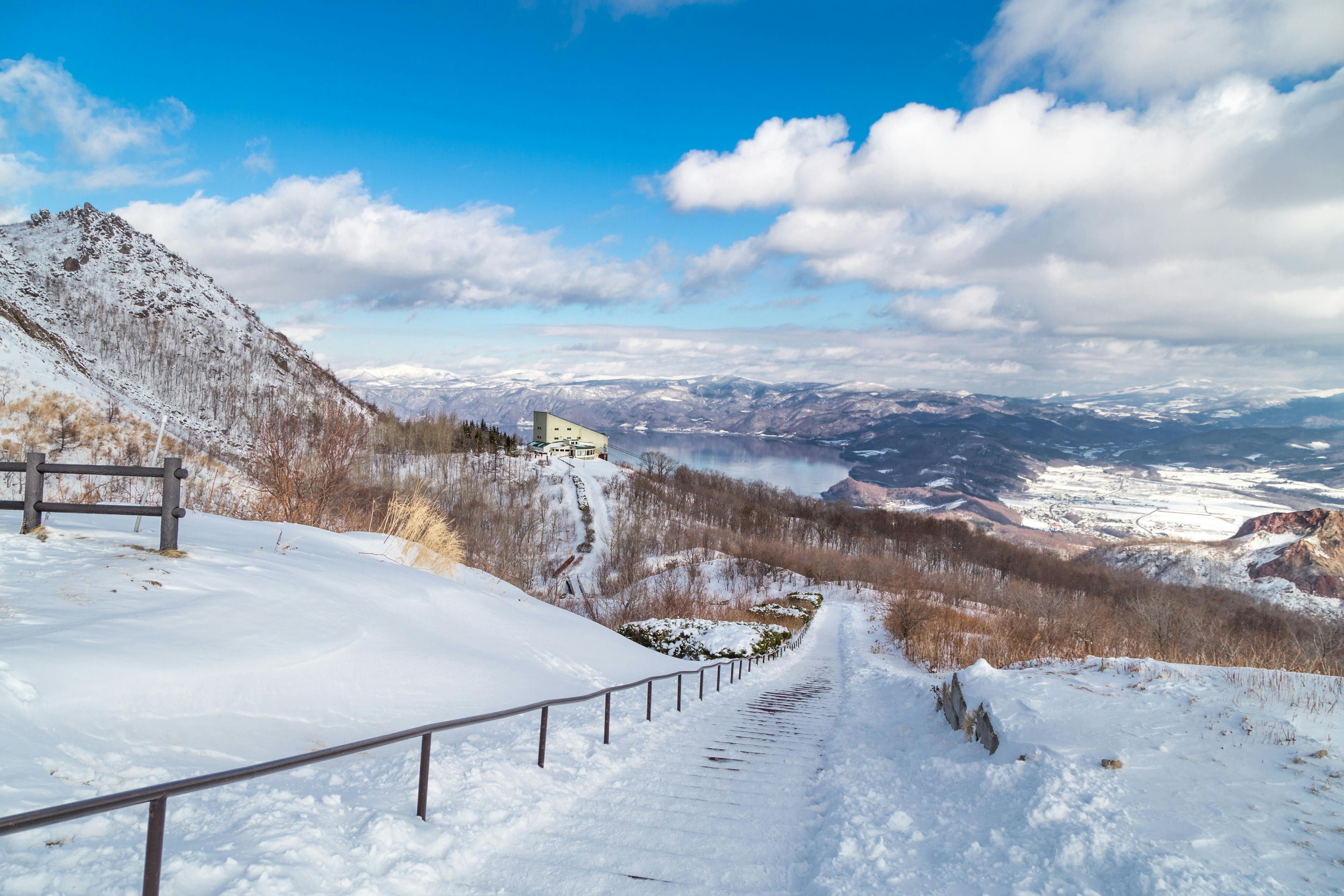 A snow-covered path with handrails descends a mountain, leading to a distant building. Snow blankets the landscape, with mountains, a lake, and a cloudy blue sky in the background.