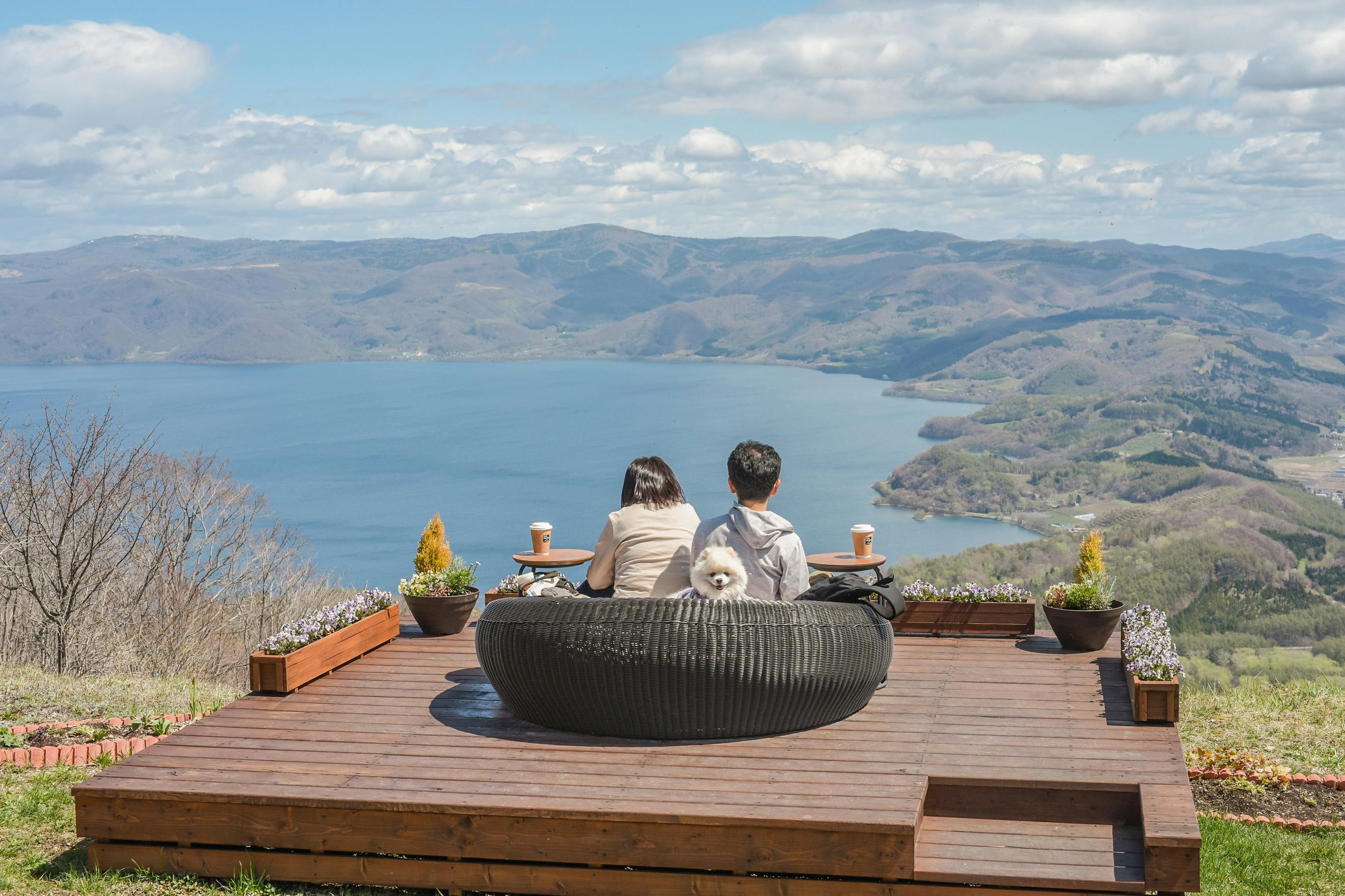 A couple sits on a round bench with a small white dog between them, enjoying drinks and a scenic view of a lake and mountains under a blue sky with scattered clouds.