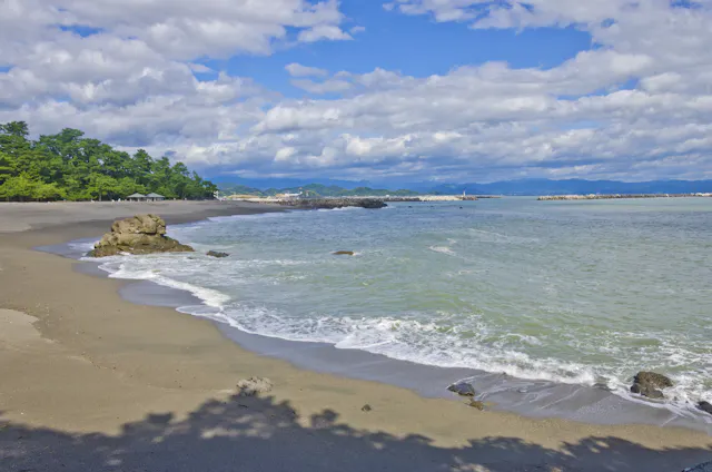 A peaceful sandy beach with scattered rocks, gentle waves, and clear blue-green water. Green trees line the shore under a partly cloudy sky with distant hills visible in the background.