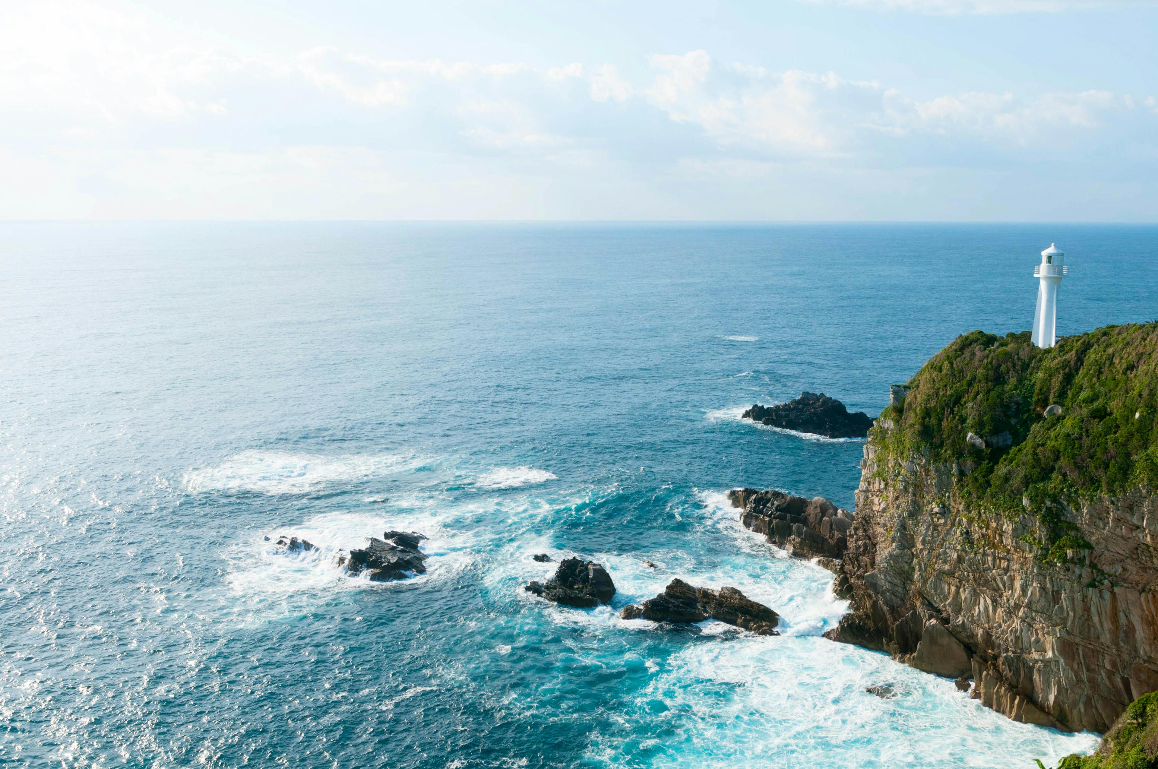 A white lighthouse stands atop a rocky, green cliff overlooking a vast, blue ocean with scattered rocks and gentle waves under a partly cloudy sky.