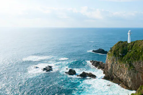 A white lighthouse stands atop a rocky, green cliff overlooking a vast, blue ocean with scattered rocks and gentle waves under a partly cloudy sky.