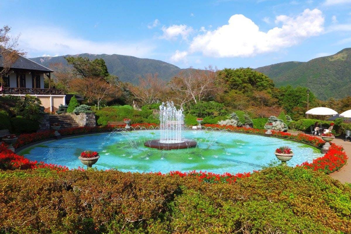 A round fountain sprays water in the center of a landscaped garden, surrounded by red flowers, green shrubs, and distant mountains under a blue sky with scattered clouds.