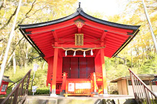 A bright red Japanese Shinto shrine with a curved roof stands among green trees. Steps lead up to the entrance, which features hanging white paper ornaments and a wooden placard with Japanese characters.