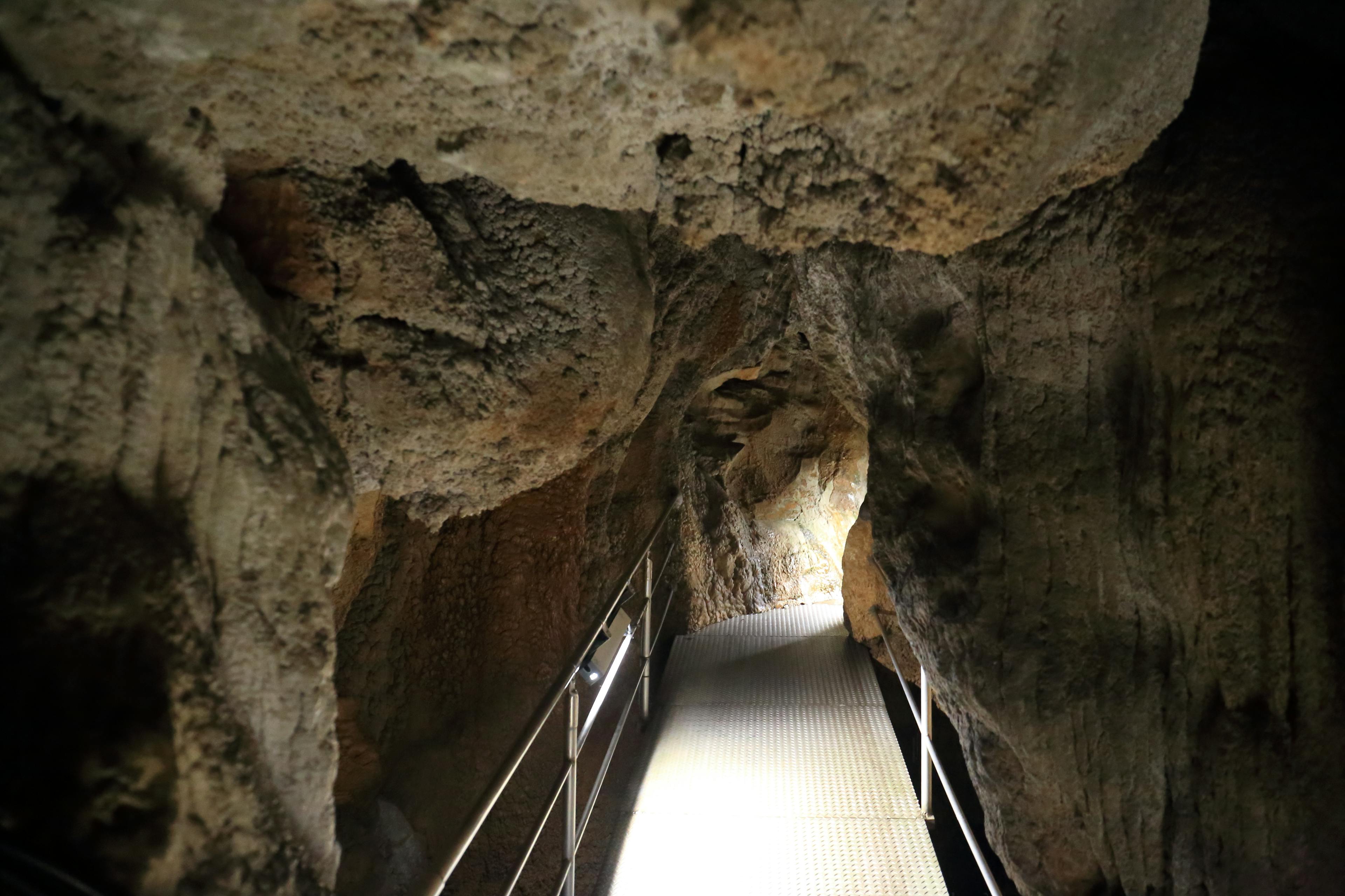 A narrow metal walkway with railings leads through a rocky cave tunnel, illuminated by soft lights along the path, with natural stone formations overhead and on the sides.