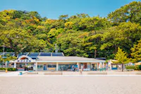 A small aquarium building with a blue sign stands in front of a forested hillside, with a gravel area and scattered visitors in the foreground under a clear, sunny sky.