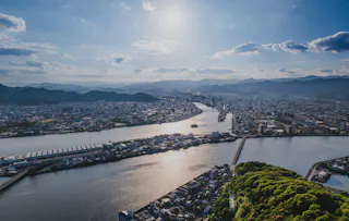 Aerial view of a city with a wide river running through it, surrounded by mountains under a bright, partly cloudy sky. The sun reflects off the water, and the foreground shows green hills and urban buildings.