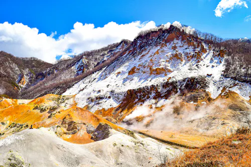 Snow-dusted mountain with orange and yellow rocky slopes, patches of steam rising from the ground, under a bright blue sky with white clouds. Sparse trees cover parts of the ridgeline.