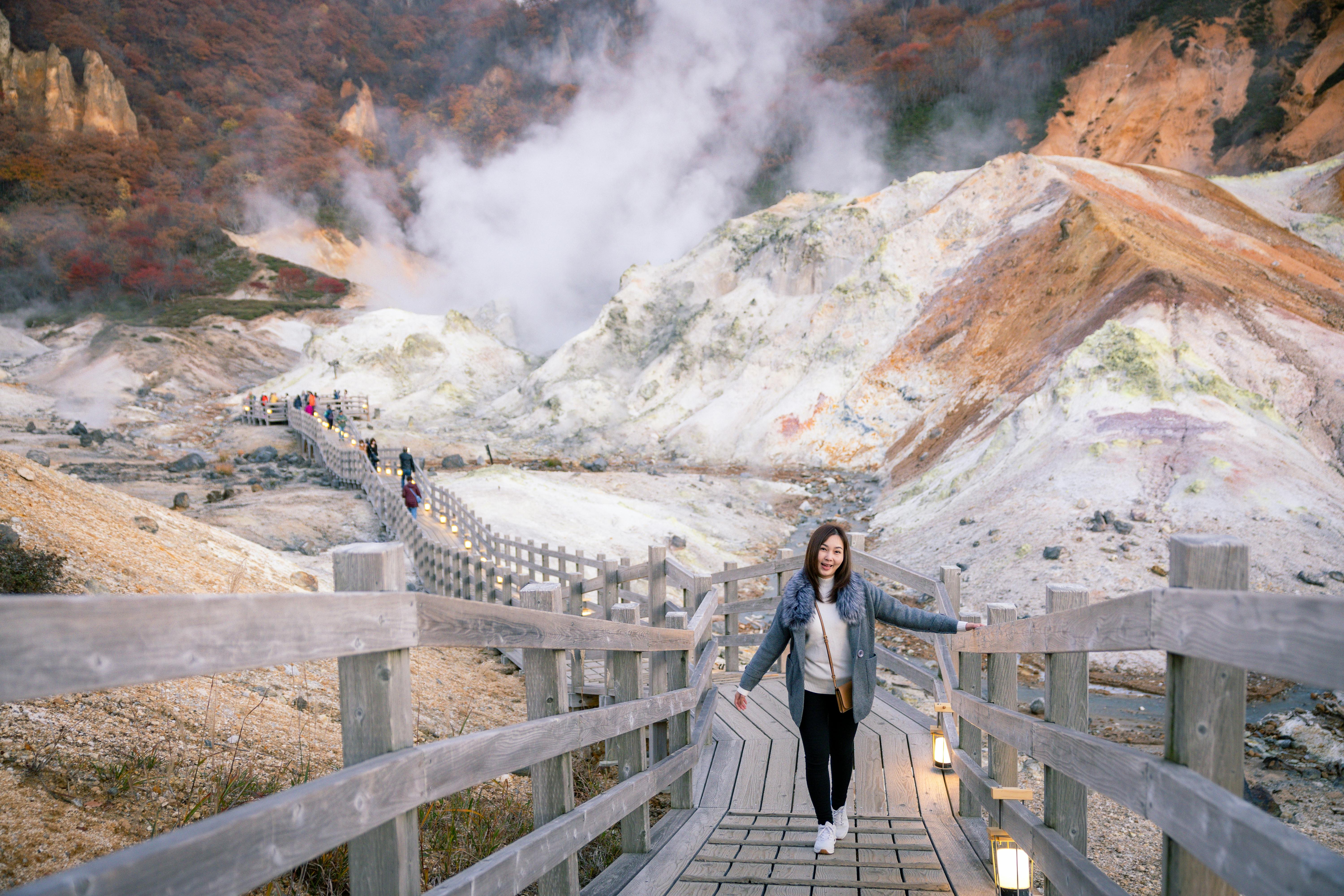 A person walks on a wooden walkway surrounded by steaming volcanic terrain and colorful hills, with more people visible in the distance. The area is natural, scenic, and mountainous.