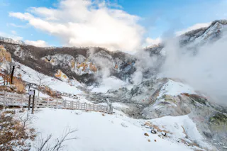 Snow-covered landscape with wooden fences and steam rising from geothermal vents, set against rugged mountains and a partly cloudy blue sky.
