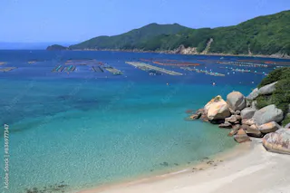 A clear blue ocean with floating fish farms, a sandy beach, large rocks, and green hills in the background under a sunny sky.