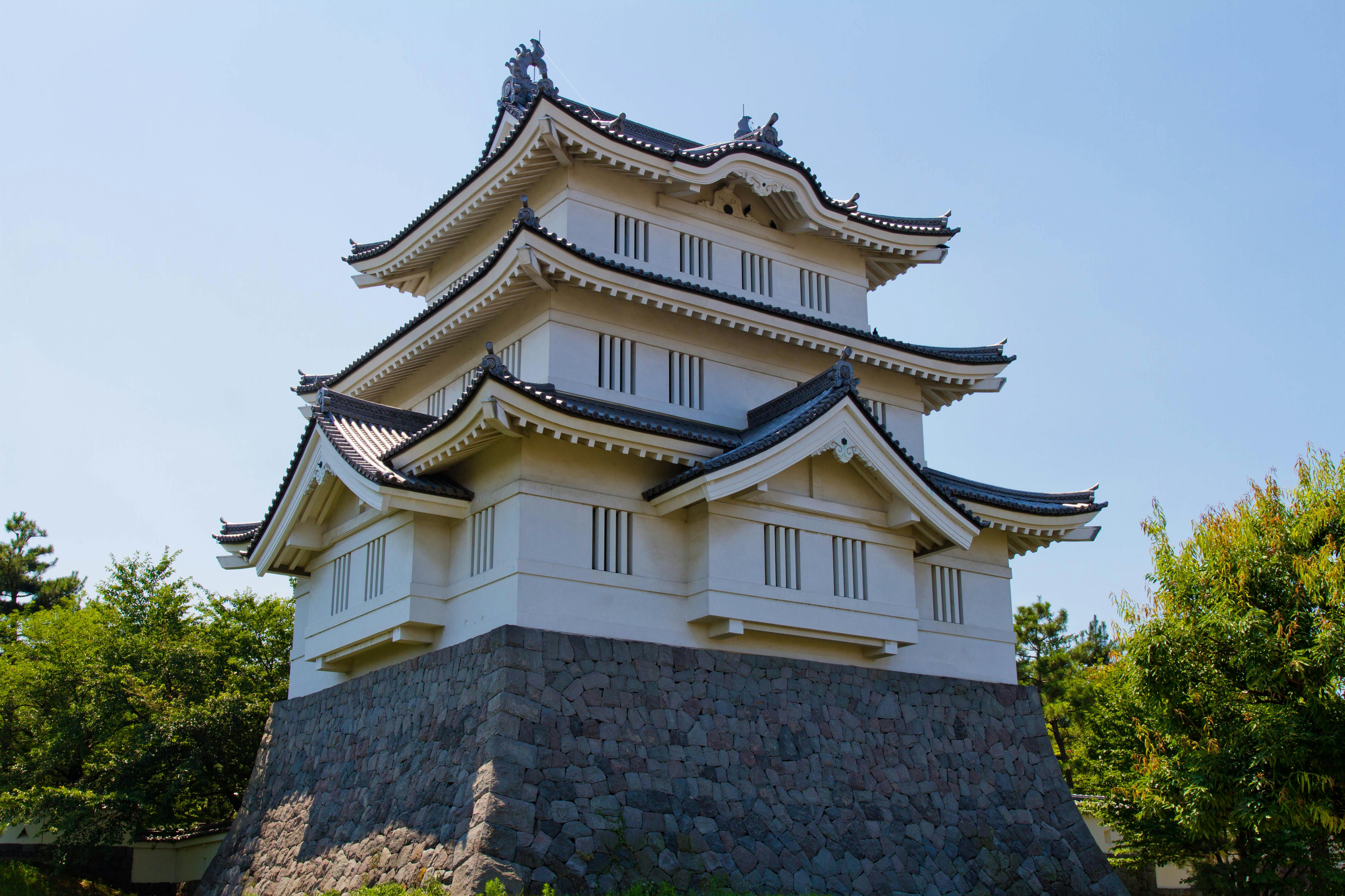 A traditional Japanese castle with white walls and black-tiled roofs stands on a stone base, surrounded by green trees under a clear blue sky.