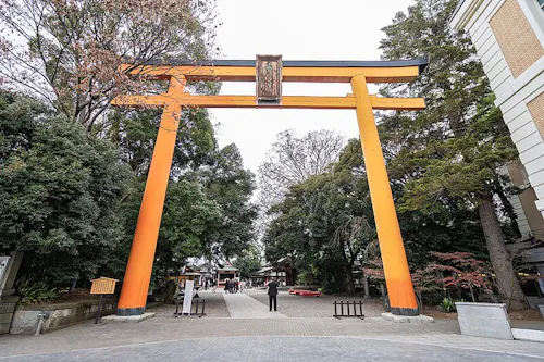 A large, bright orange torii gate stands at the entrance to a shrine, surrounded by tall trees and buildings, with a few people walking beneath it on a paved path.