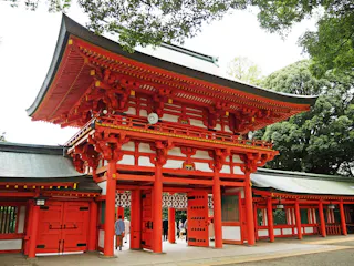 A large, traditional Japanese torii gate and entrance painted bright red with white accents, featuring ornate wooden architecture, stands surrounded by lush green trees.