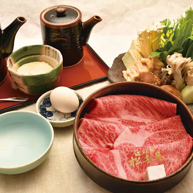 Matsukiya Cooking Class A beautifully arranged Japanese hot pot meal with marbled beef slices, mushrooms, leafy greens, an egg, dipping sauces, and ceramic pots, all set on a light-colored tablecloth.