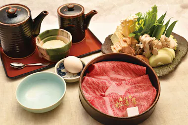 A beautifully arranged Japanese hot pot meal with marbled beef slices, mushrooms, leafy greens, an egg, dipping sauces, and ceramic pots, all set on a light-colored tablecloth.