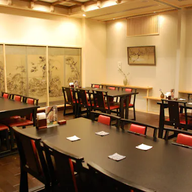 Matsukiya Cooking Class A modern Japanese restaurant dining room with dark wooden tables and chairs featuring red cushions, soft lighting, and traditional artwork on the walls. Each table has coasters and simple decorations.