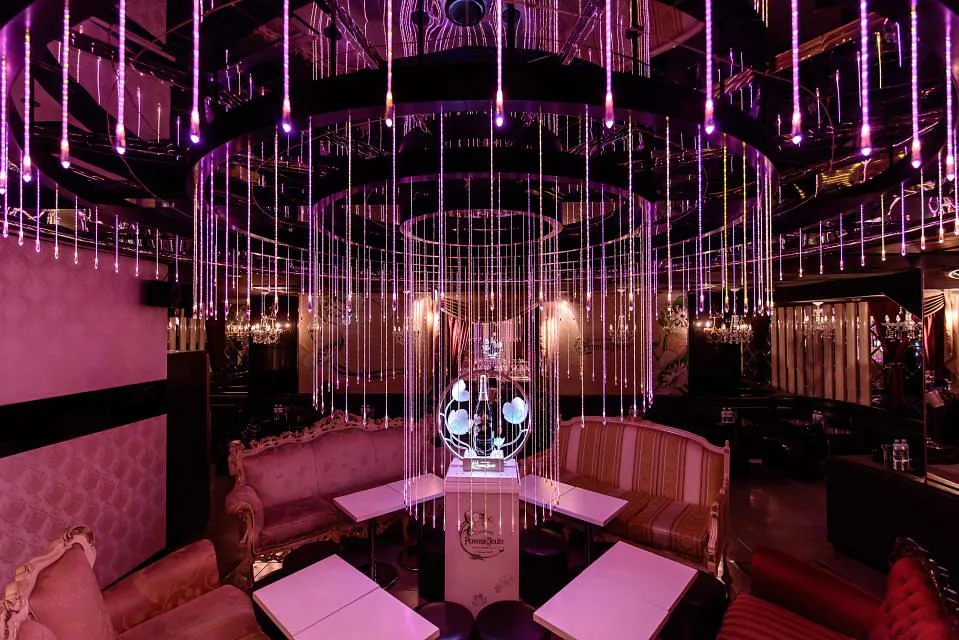 A dimly lit and opulent lounge with pink and purple neon lights, featuring plush, elegant seating. The ceiling is adorned with hanging LED strands, creating a sparkling effect. The central focus is a modern, transparent DJ booth. The ambiance is luxurious and sophisticated.