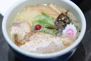 A bowl of ramen with sliced pork, sesame seeds, bamboo shoots, green onions, a pink-and-white fish cake, a red pickled plum, and dark mushrooms in a creamy broth.