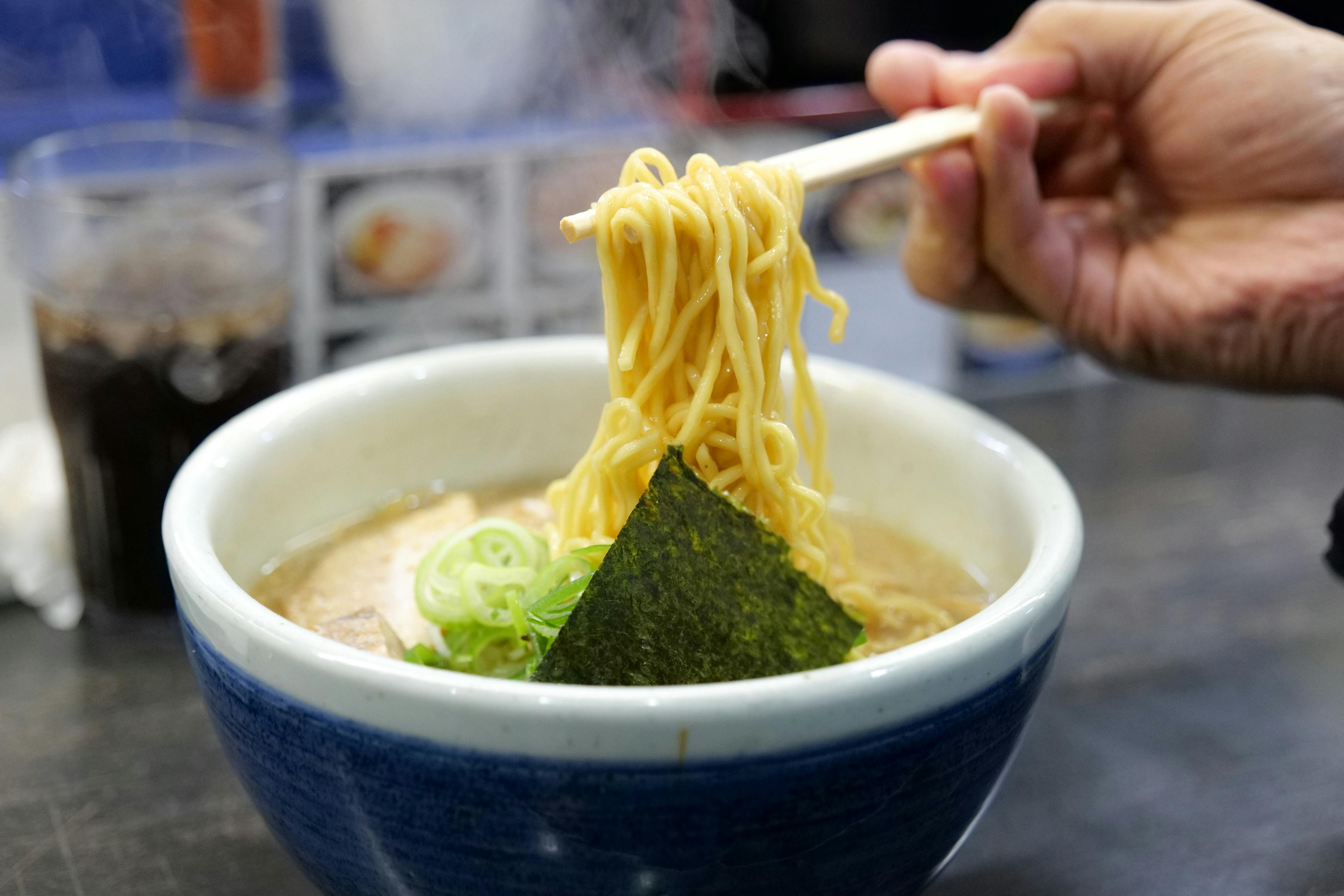 A hand holds chopsticks lifting noodles from a steaming bowl of ramen topped with seaweed, green onions, and broth. A glass of dark beverage sits in the background.