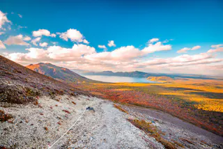 A scenic view from a mountain slope shows a rocky path, colorful autumn trees below, a large lake, distant mountains, and a bright sky with scattered clouds.