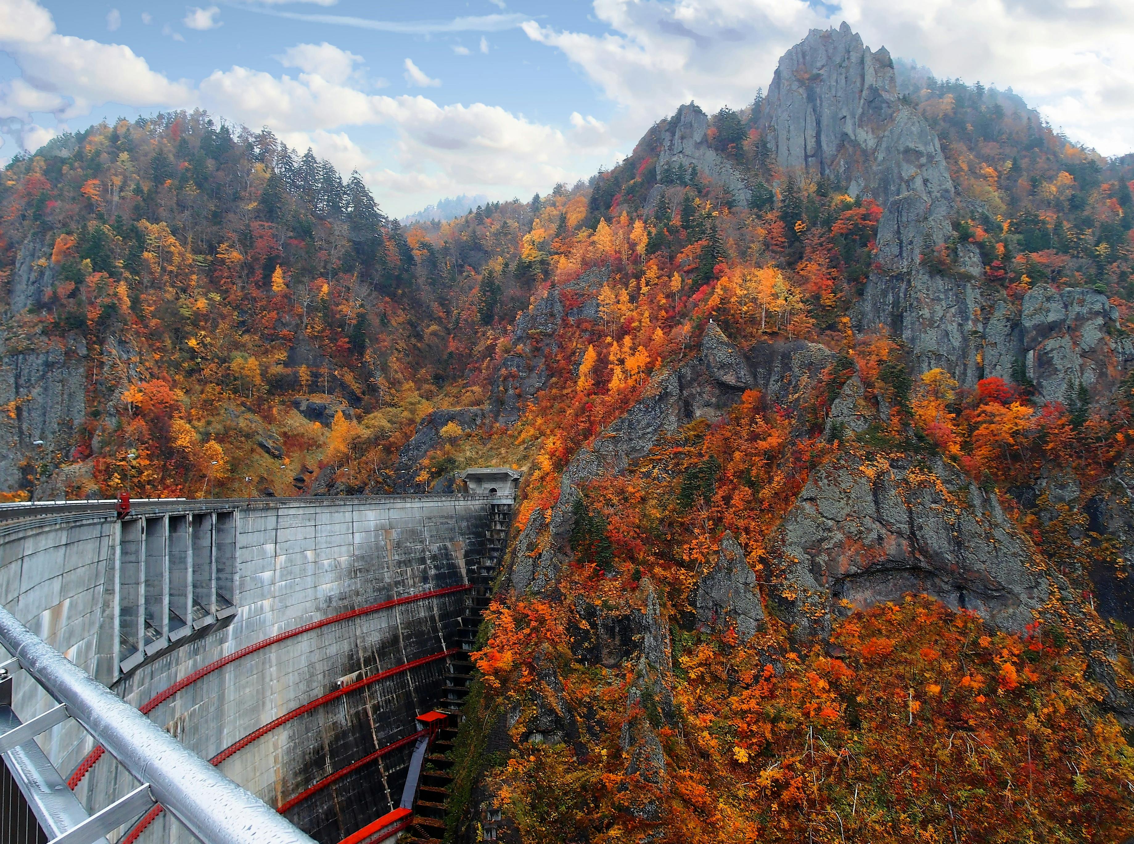 A curved concrete dam sits among rugged, rocky mountains covered in vibrant autumn foliage, with trees displaying red, orange, and yellow leaves under a partly cloudy sky.