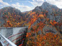 A curved concrete dam sits among rugged, rocky mountains covered in vibrant autumn foliage, with trees displaying red, orange, and yellow leaves under a partly cloudy sky.