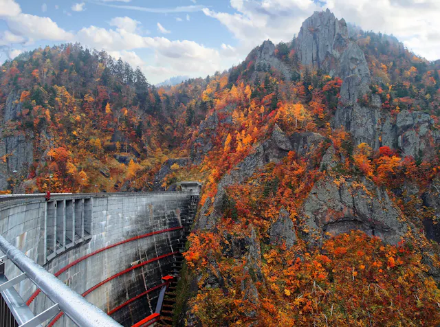 A curved concrete dam sits among rugged, rocky mountains covered in vibrant autumn foliage, with trees displaying red, orange, and yellow leaves under a partly cloudy sky.