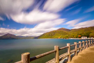 A scenic lakeside view with turquoise water, a wooden fence in the foreground, green hills and mountains in the background, and a dramatic sky with streaked white clouds from long exposure.