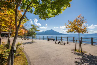 A scenic lakeside walkway with benches, bordered by green and orange autumn trees. In the background, a mountain rises under a clear blue sky with a few clouds.