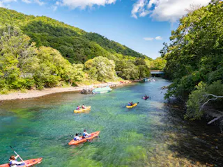 People kayaking on a clear, blue-green river surrounded by lush green trees and hills under a bright, partly cloudy sky. Several small boats are docked along the riverbank.