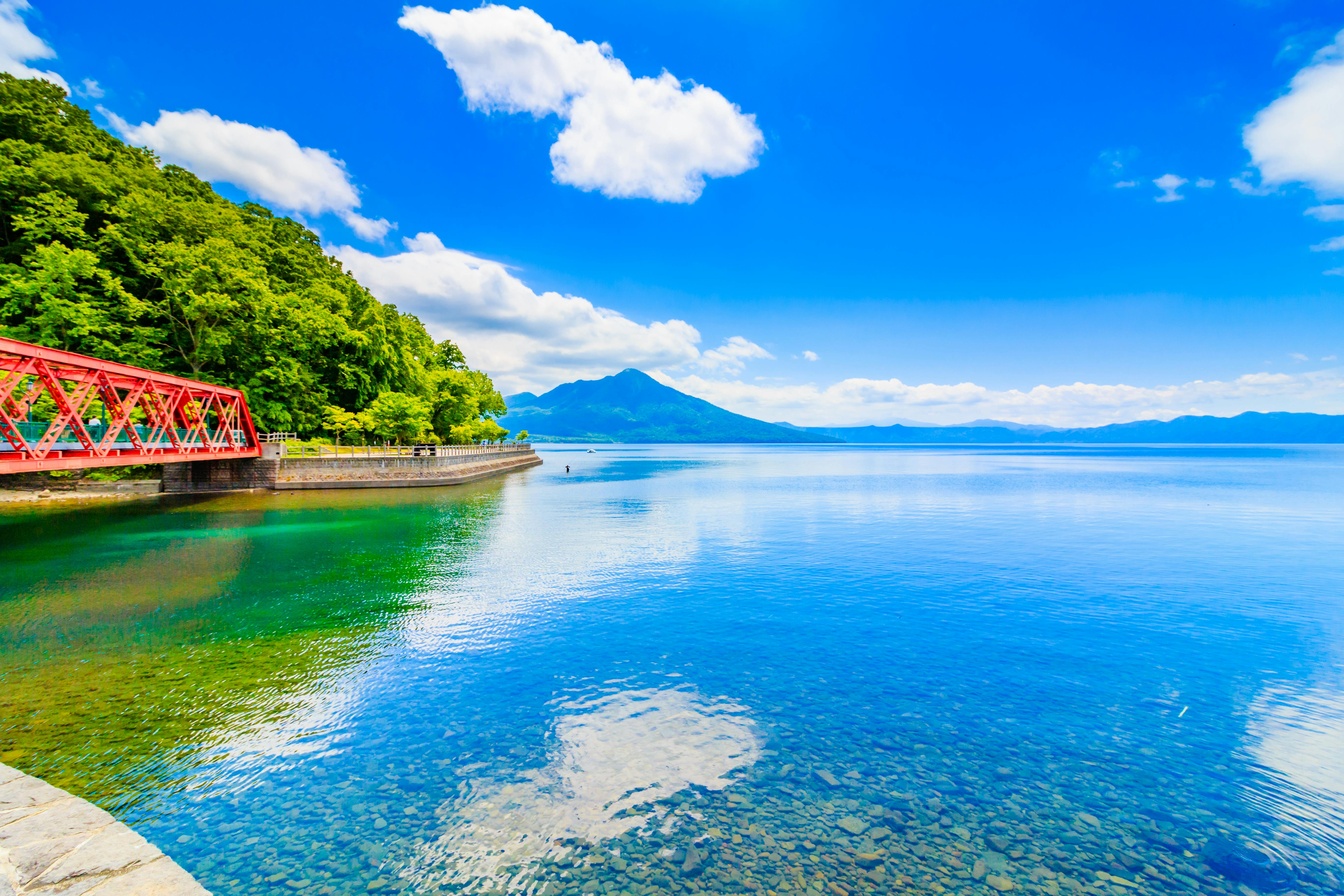 A clear blue lake with a red bridge and a tree-lined shore on the left, distant mountains under a bright blue sky with scattered clouds reflected in the calm water.