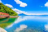 A clear blue lake with a red bridge and a tree-lined shore on the left, distant mountains under a bright blue sky with scattered clouds reflected in the calm water.