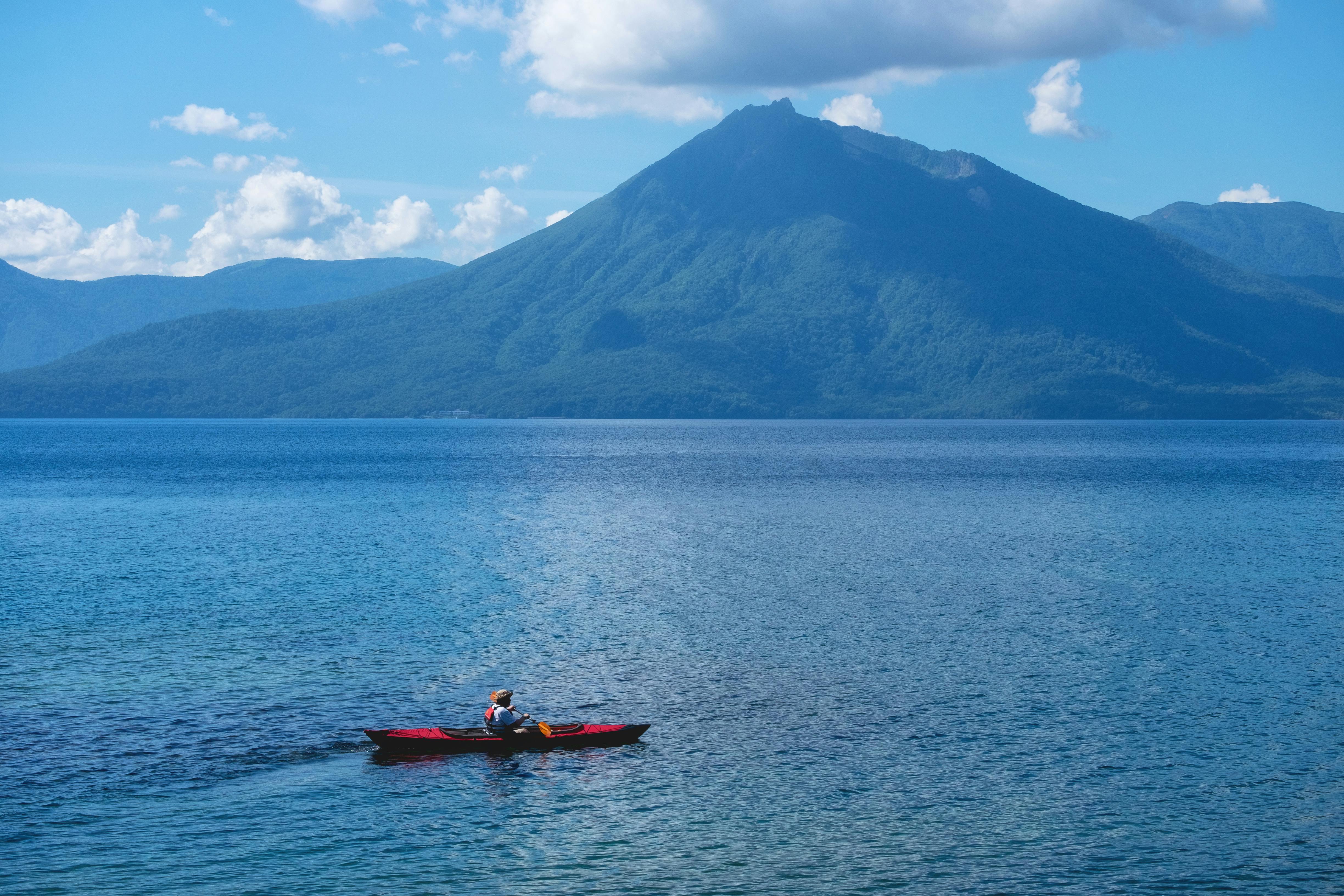A person in a red kayak paddles across a calm blue lake with a large, forested mountain and partly cloudy sky in the background.