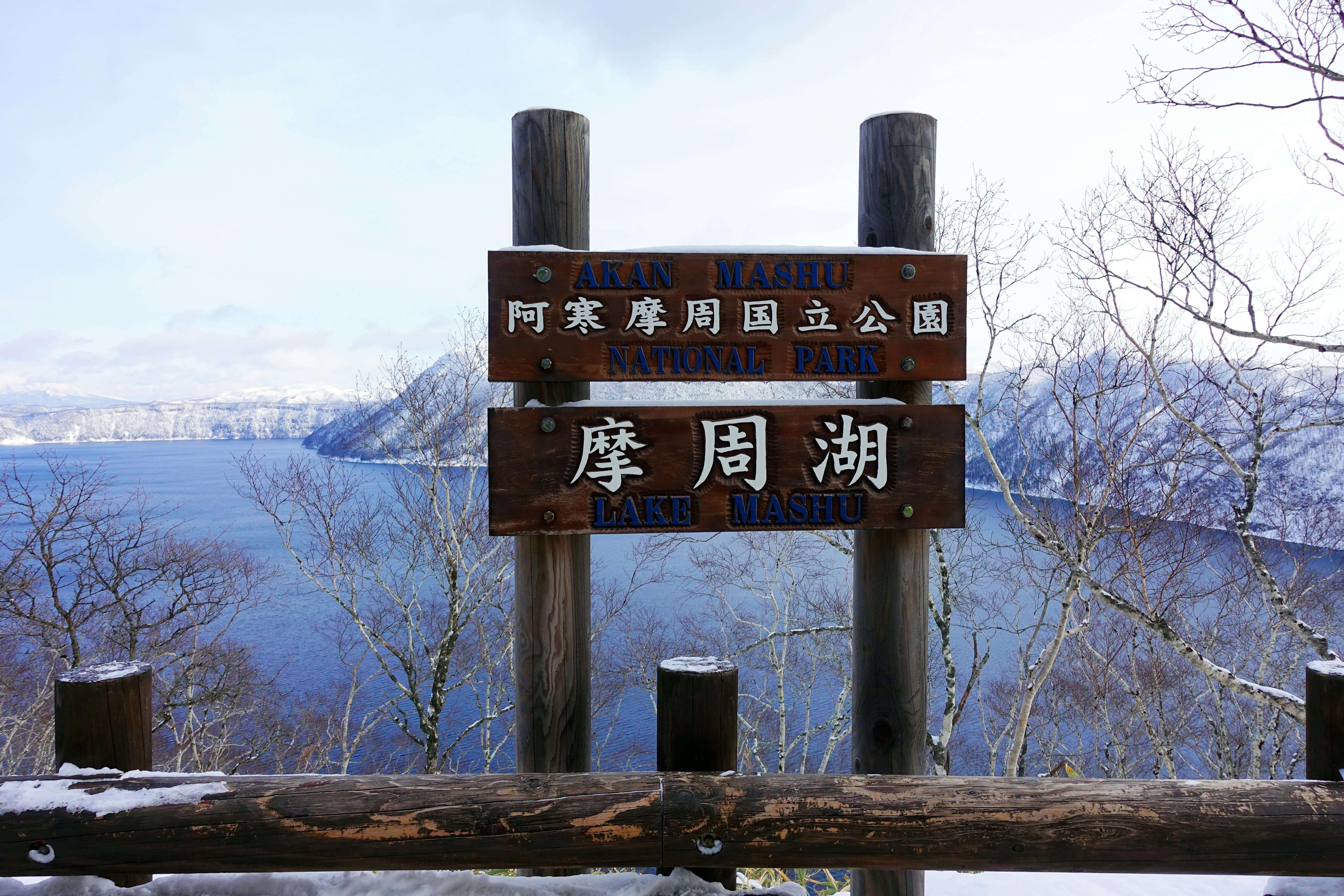Wooden signpost in front of a lake with mountains in the background, reading “Akan Mashu National Park” and “Lake Mashu” in English and Japanese; snow covers the trees and ground.