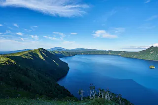 A deep blue lake surrounded by lush green hills under a bright blue sky with scattered clouds. A small island is visible in the water near the right edge of the image.