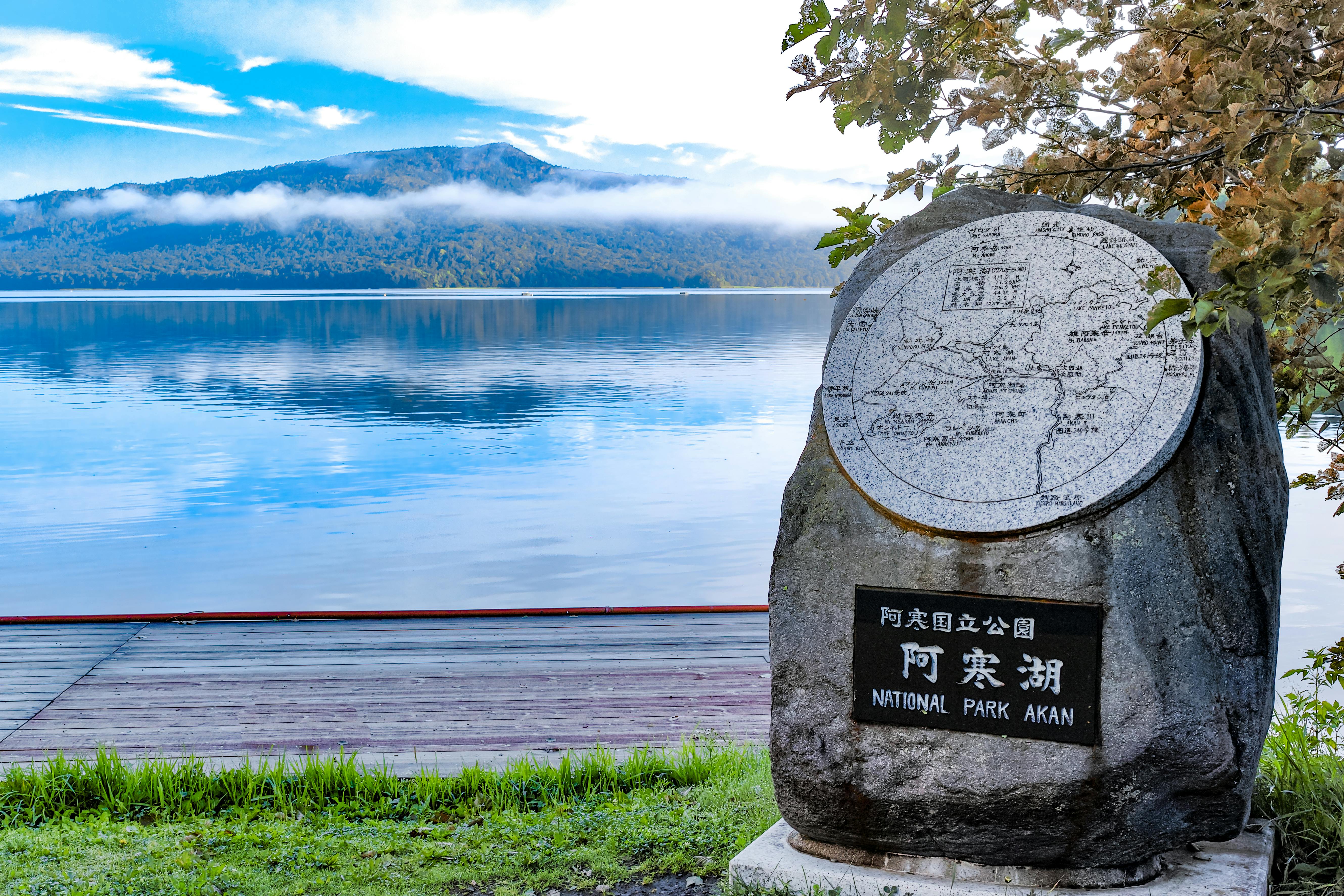 A large stone monument with a map and Japanese writing marks Akan National Park beside a calm lake, with mountains and low clouds reflected in the water under a blue sky.