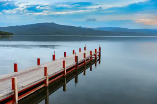 A wooden dock with red posts extends over calm, reflective water, with distant green hills and a partly cloudy blue sky in the background.
