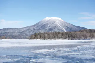 Snow-capped mountain rises behind a forest and a partially frozen lake, with patches of ice on the water under a clear blue sky.