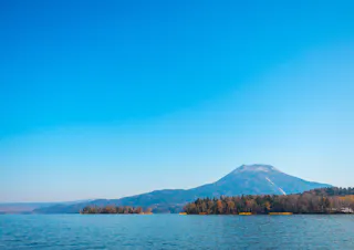 A calm lake bordered by trees with a distant mountain under a clear blue sky. The landscape is peaceful and bright, with sunlight illuminating the water and trees.