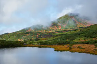 A scenic mountain landscape with green and orange foliage, partially covered by mist, reflected in a calm lake in the foreground. Low clouds and blue sky add a serene atmosphere.