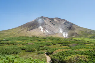 A distant volcanic mountain with patches of green vegetation at its base and slopes. White steam rises from several vents partway up the mountain under a clear blue sky.