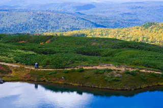Two hikers walk along a narrow path beside a calm blue lake, surrounded by lush green hills and distant forested mountains under a bright sky.