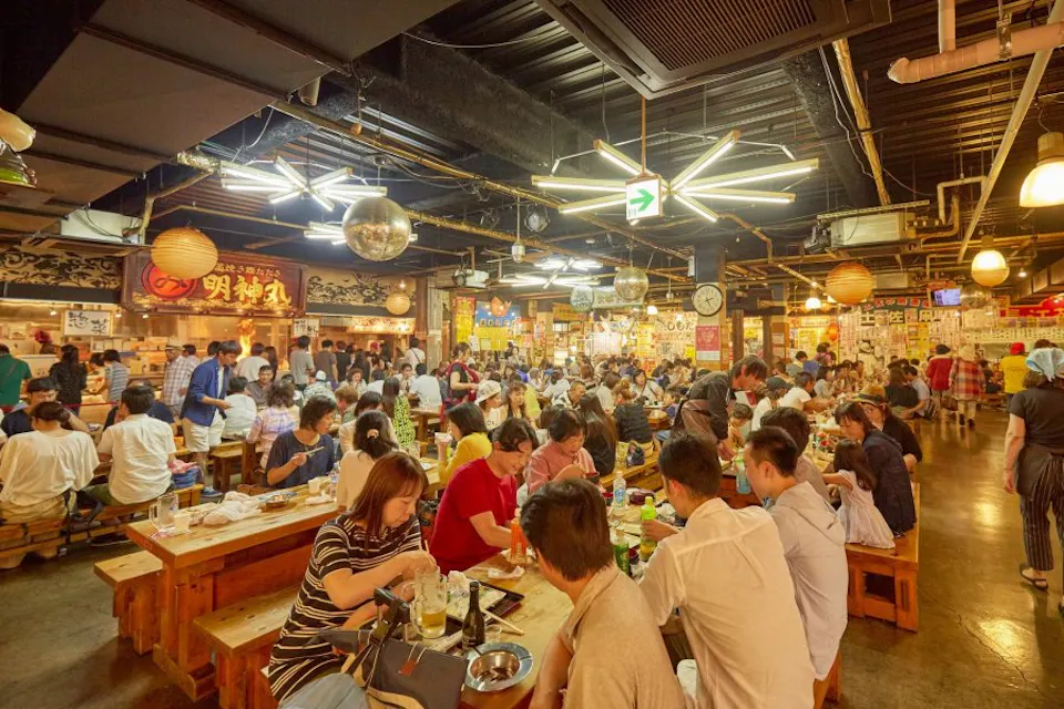 A busy indoor food market with wooden tables packed with people eating and socializing. The warm lighting and colorful signs create a lively atmosphere. Various food stalls can be seen in the background, with customers waiting in line.