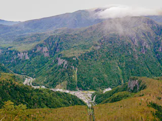 A lush, green mountainous landscape with a valley below containing winding roads and a small settlement, shrouded in light mist and clouds near the mountain peaks.