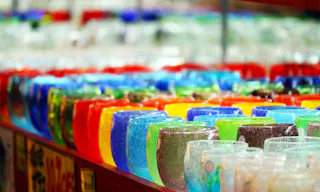 Rows of colorful glass cups are neatly arranged on a display table, creating a vibrant and eye-catching pattern with various shades of blue, yellow, red, green, and clear glass.