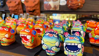 Rows of colorful, smiling Shisa lion-dog figurines are displayed on a shelf, each with bright patterns and joyful expressions. Blurred traditional masks hang in the background, adding to the lively atmosphere.