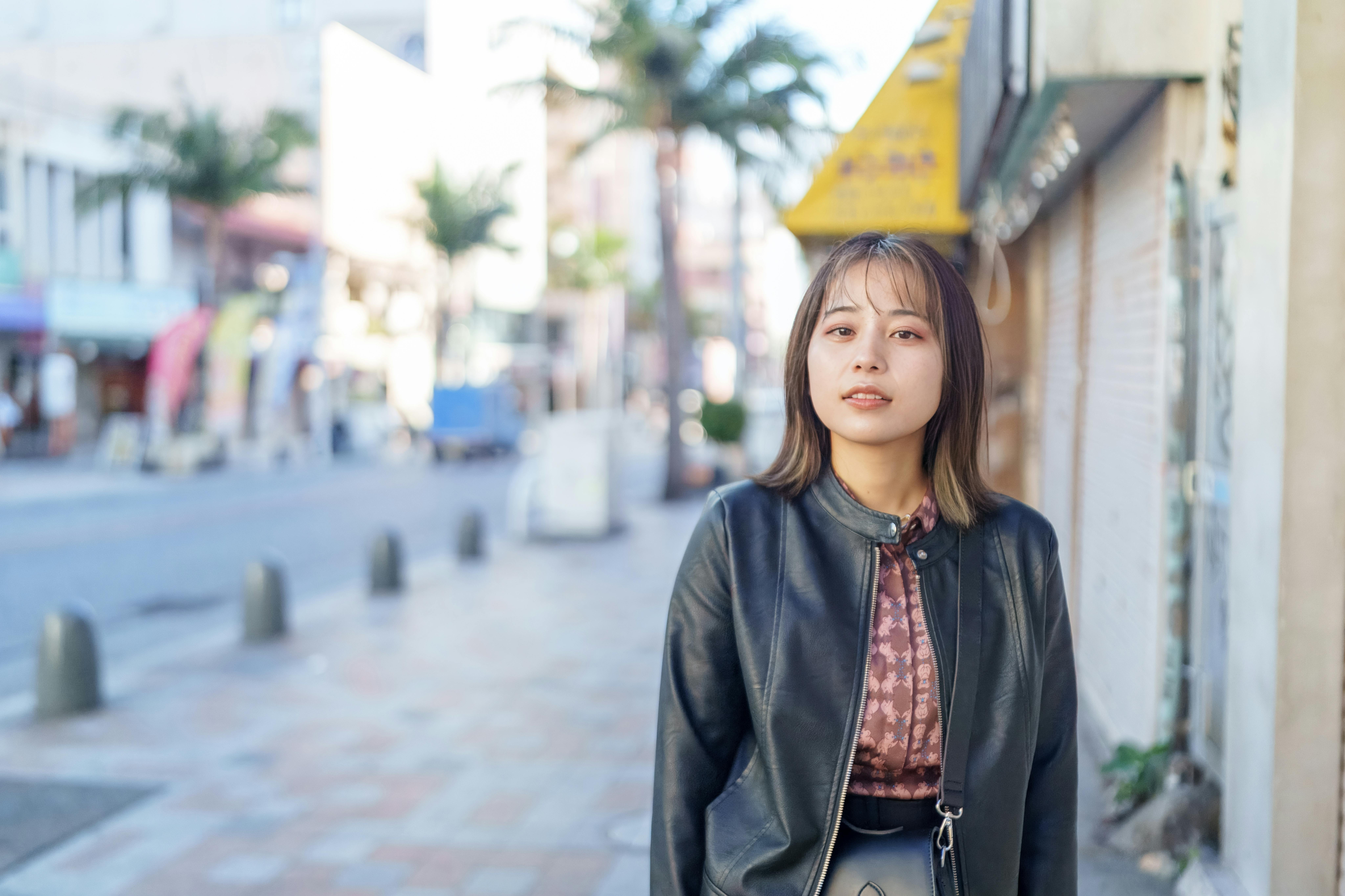 A young woman with shoulder-length hair wearing a black leather jacket stands on a sunlit city street with palm trees and shops in the background.