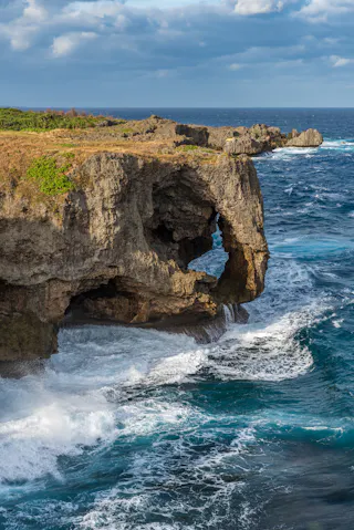 Rocky cliff with a natural arch formation overlooks rough, blue ocean waves crashing against the shore under a partly cloudy sky. Sparse grass and vegetation cover the top of the cliff.