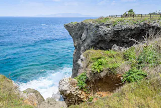 A rocky cliff covered in green plants overlooks bright blue ocean waves. A wooden fence runs along the top right edge, and distant mountains are visible on the horizon under a partly cloudy sky.